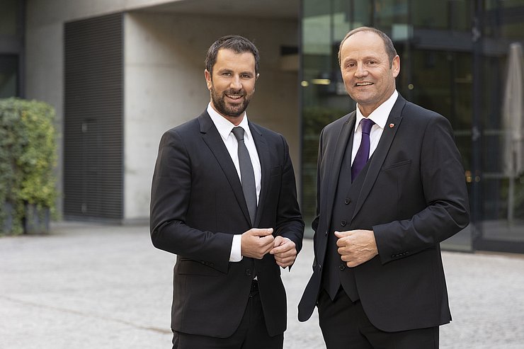 © Land Tirol/Die Fotografen Dornauer und Geisler stehen in dunklen Anzügen nebeneinander und lächen in die Kamera