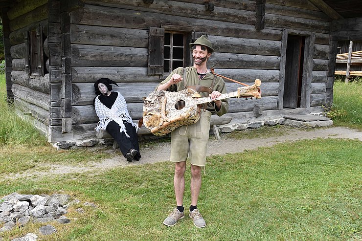 © Land Tirol/Feuersinger Almajuri hat einen Hut auf und trägt einen Bart. Er spielt auf einer Gitarre vor einer Hütte. Hinter ihm sitzt ein Fabelwesen auf einer Bank vor der Hütte.