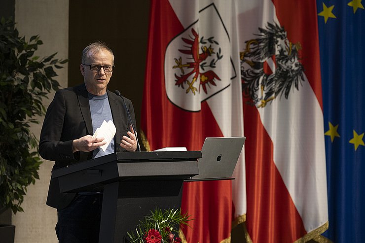 © Land Tirol/Die Fotografen Ein Mann an einem Rednerpult gestikuliert und spricht zum Publikum.