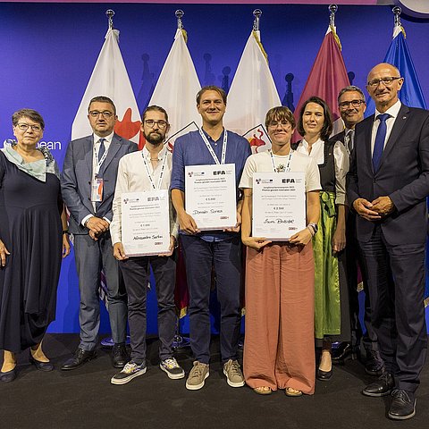 Mehrere Personen stehen vor den Flaggen der Euregio, die mittleren drei halten Urkunden.