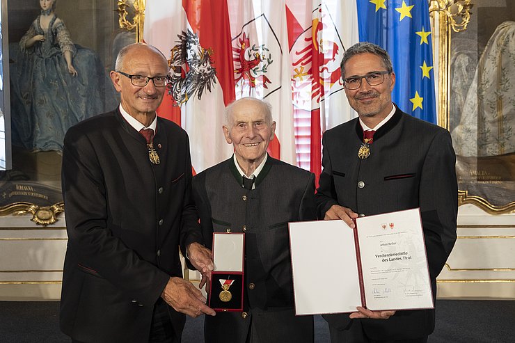 © Land Tirol/Die Fotografen links LH Mattle, in der Mitte Herr Keller und rechts LH Kompatscher, halten Medaille und Verdiensturkunde