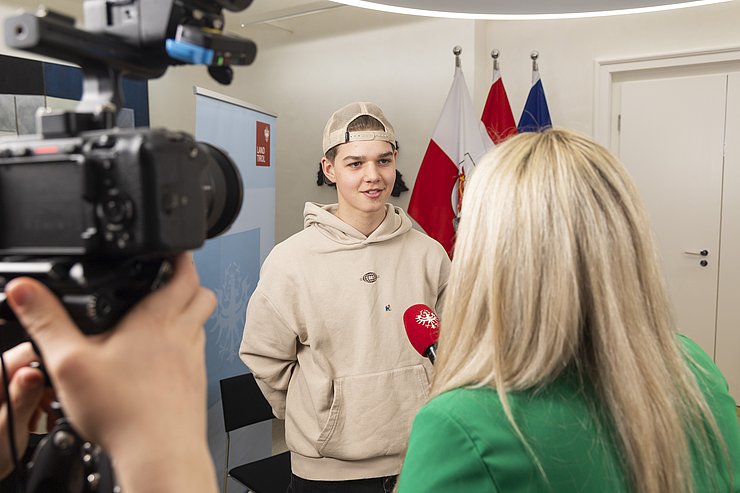 © Land Tirol/Die Fotografen Ein Schüler steht vor einer Kamera, von einer Dame wird ihm ein Mikrofon vorgehalten
