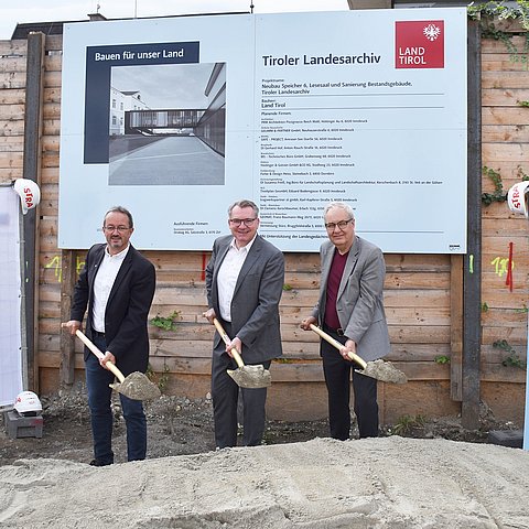 Foto vom Spatenstich; drei Personen mit Schaufeln voll Sand vor Sandhaufen; dahinter Bautafel
