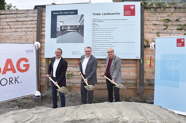 © Land Tirol/Pölzl Foto vom Spatenstich; drei Personen mit Schaufeln voll Sand vor Sandhaufen; dahinter Bautafel