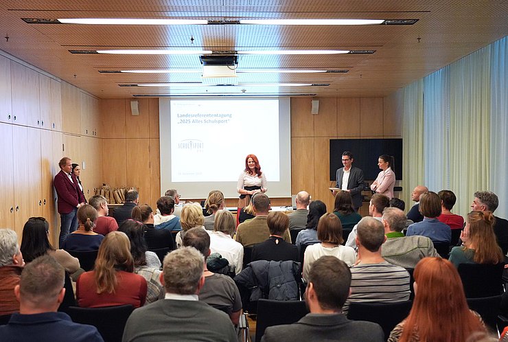 Aufnahme eines gut gefüllten Konferenzsaals, in dem eine Frau vor einer Leinwand spricht. 