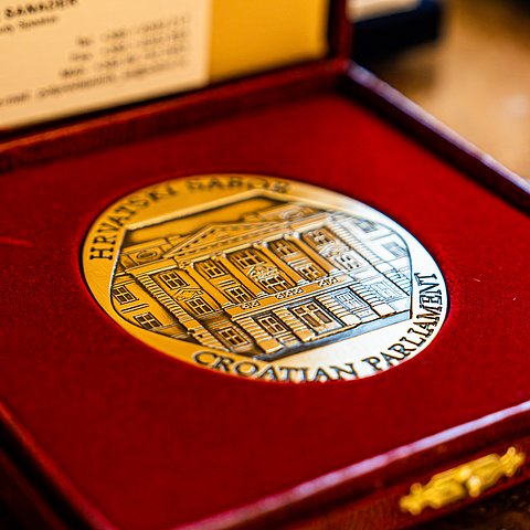 Schachtel mit rotem Samt. Darin liegt eine goldene Medaille. Auf dieser ist die Hausfront des kroatischen Parlaments eingraviert. Zudem der Schriftzug: Croatian Parliament und Hrvatski Sabor